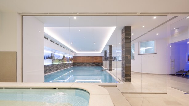 Ein moderner, überdachter Schwimmbereich mit großen Glaswänden, einer Holzwand und einer Rezeption mit blau beleuchtetem Empfangstresen.