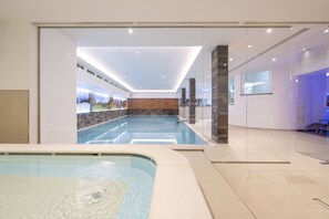A modern indoor swimming pool with clear glass walls, a wooden wall, and a reception area with a blue-lit desk. - Astoria (Canazei)