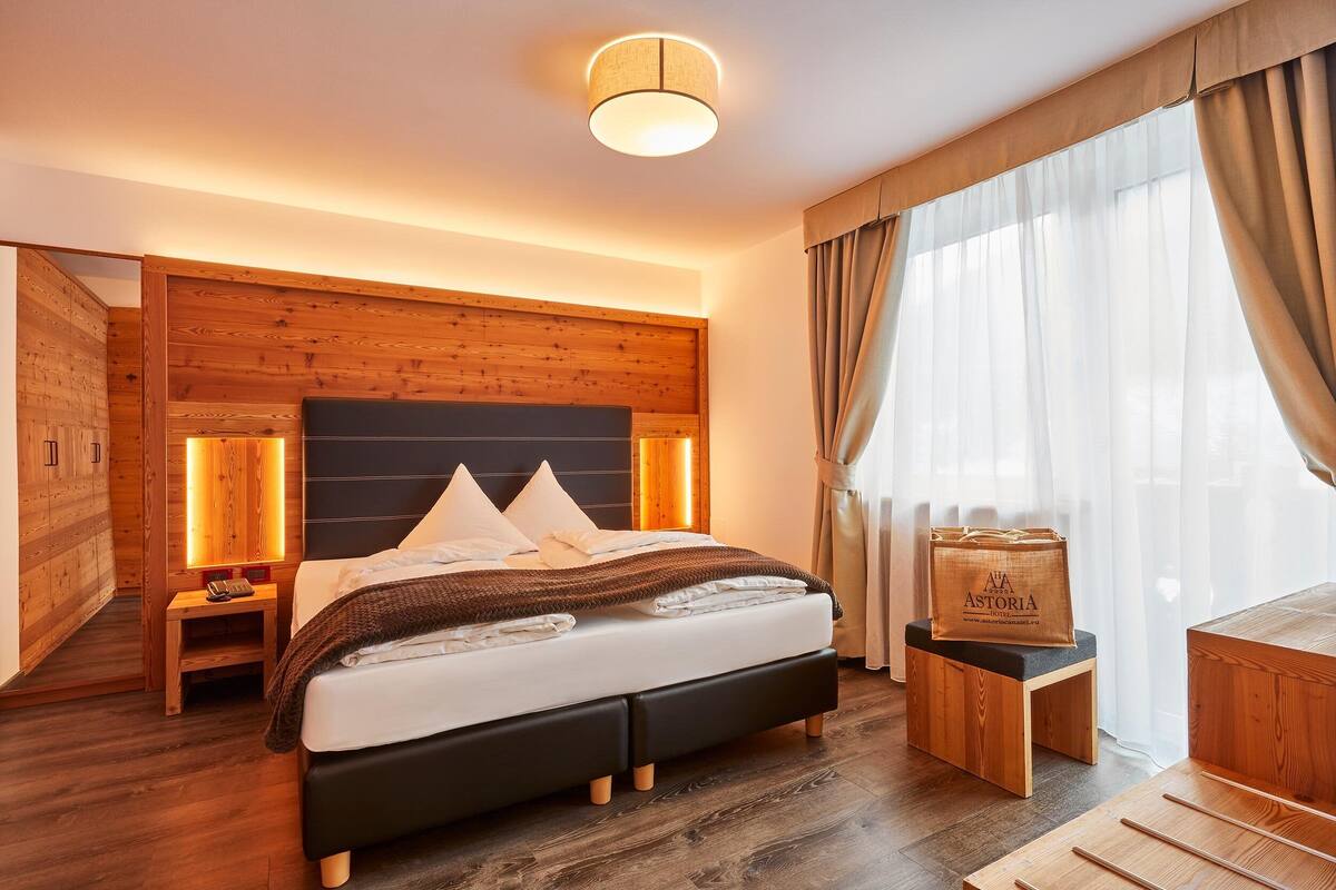 Una habitación de hotel moderna con cabecera de madera, cama con ropa blanca, mesita de noche con lámpara y una ventana grande con cortinas.