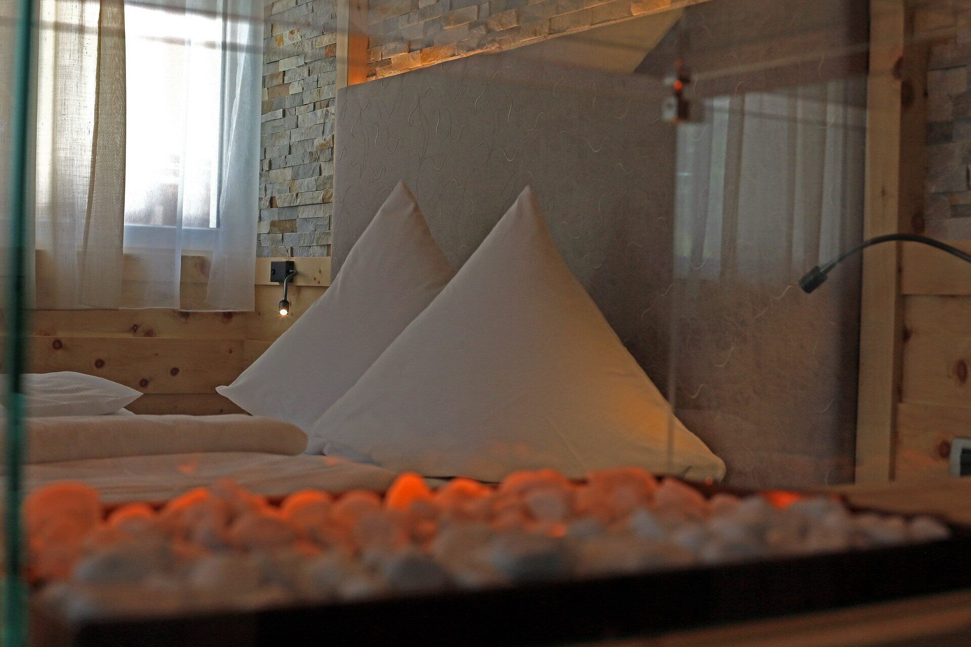 A bed with white pillows and a wooden headboard in a room with a stone wall and a window with curtains.