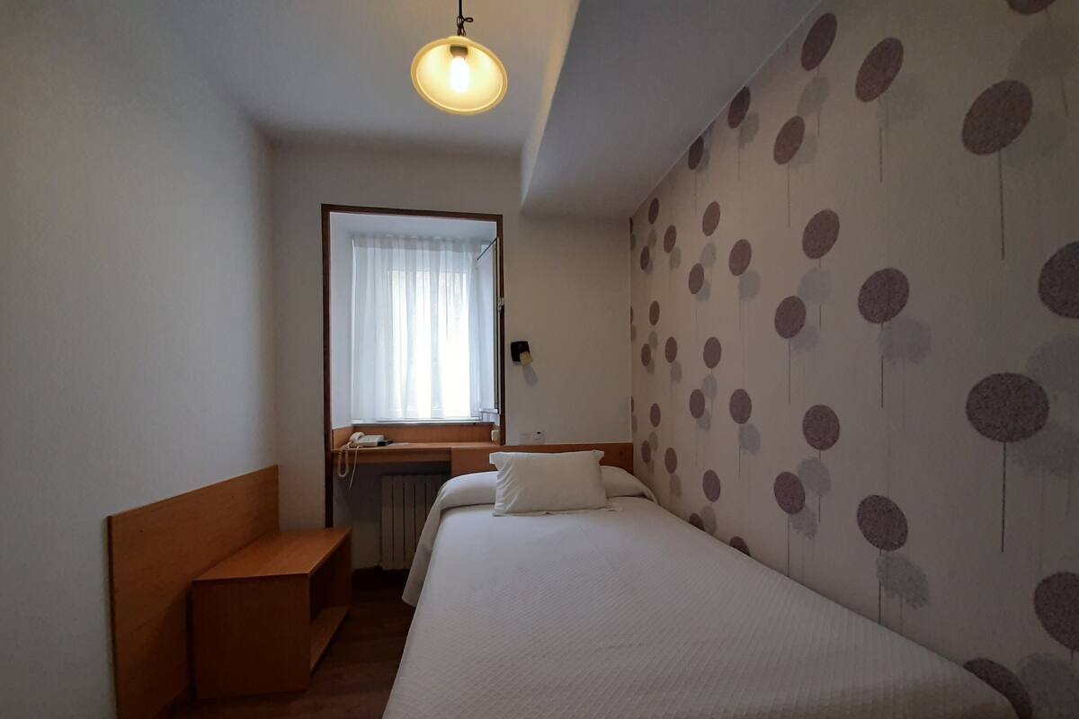 A hotel room with a bed, a desk with a telephone, a window with curtains, and a wall with a polka dot pattern.
