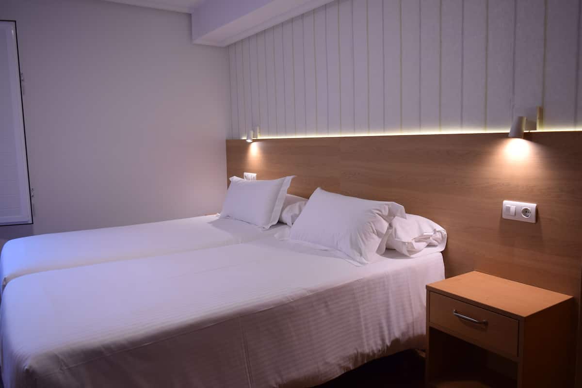 A neatly made bed with white linens and pillows, a wooden nightstand with a drawer, and a wall-mounted light fixture.