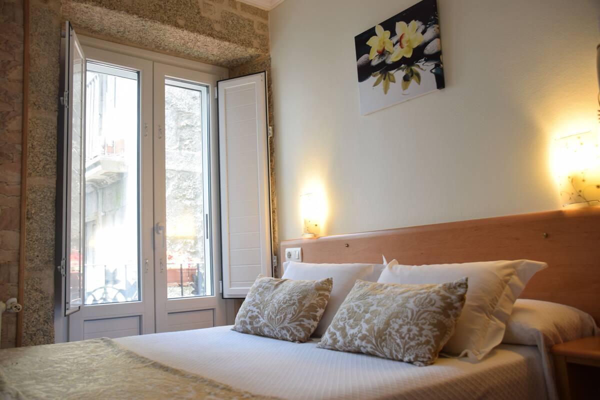A bedroom with a large bed, two pillows, a nightstand, a wall-mounted lamp, and a window with a view of the outside.