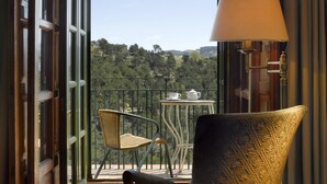 A balcony with a chair and a table, a lamp, and a view of a forested area.