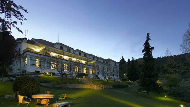 A multi-story hotel with a well-lit exterior, a grassy courtyard with outdoor seating, and a forested hillside in the background.