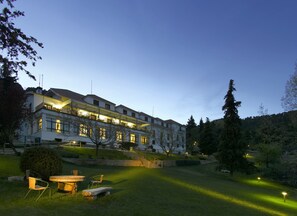 A multi-story hotel with a well-lit exterior, a grassy courtyard with outdoor seating, and a forested hillside in the background. - Parador De Cazorla (Cazorla)
