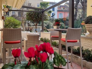 A covered outdoor seating area with wicker furniture and red cushions, surrounded by potted plants and a view of buildings. - Charly Hotel (Milan)