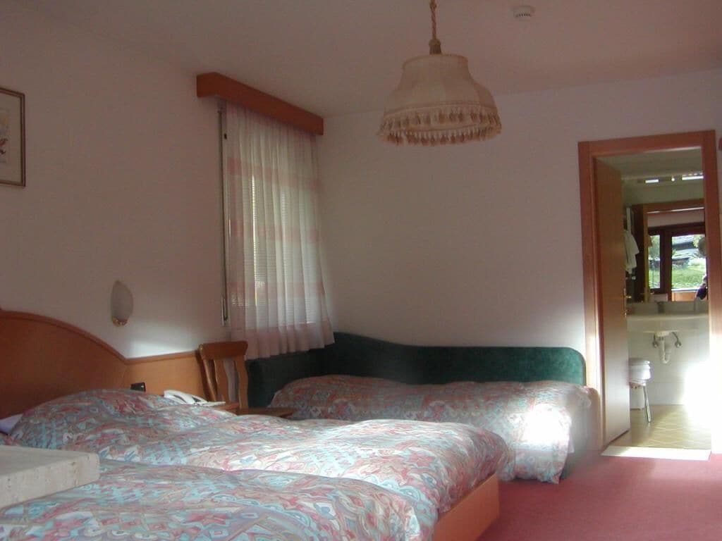 A bedroom with two beds, a wooden headboard, a hanging light fixture, a window with curtains, and a view of a bathroom.