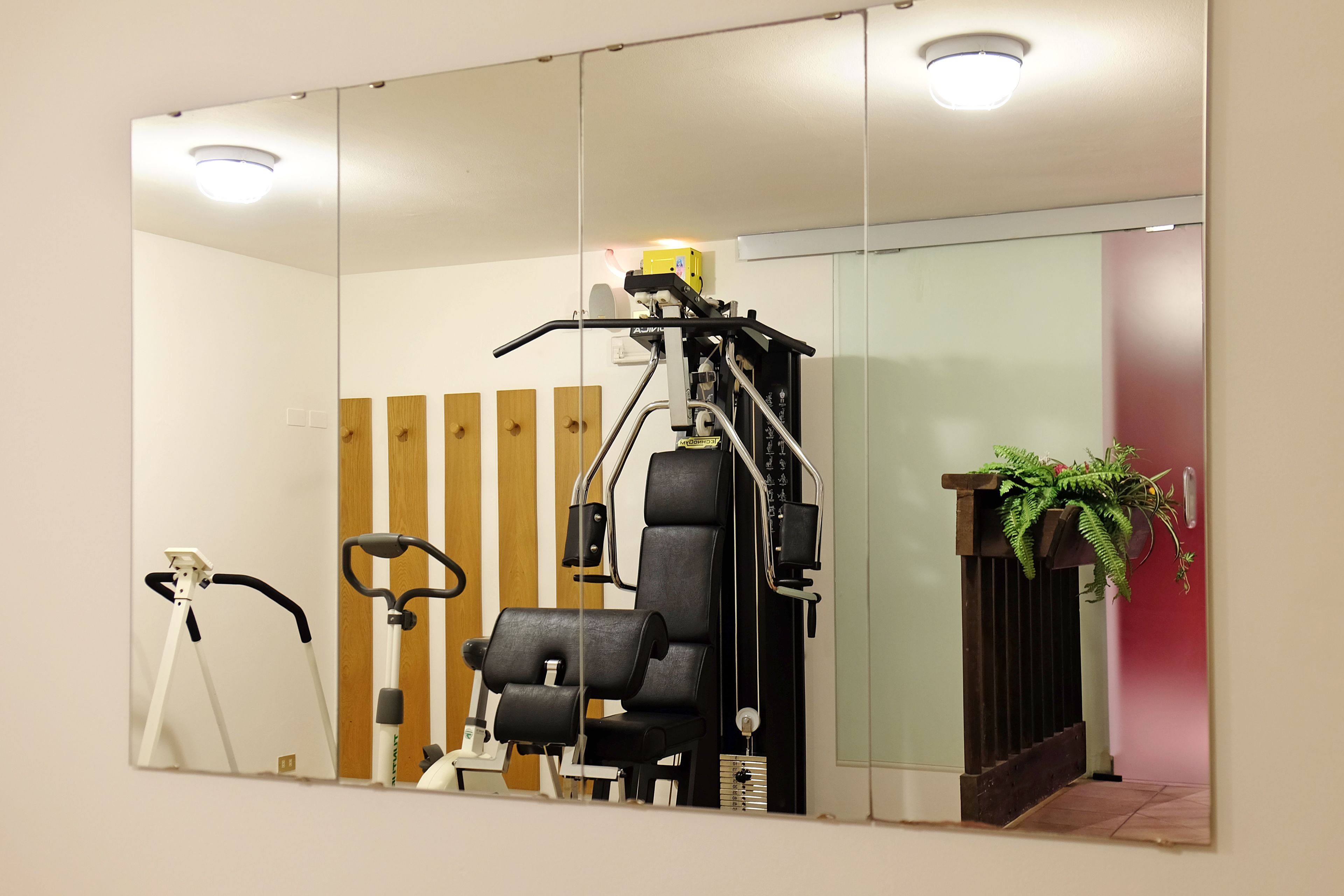 A mirrored wall reflecting a gym area with exercise equipment, a standing bike, and a potted plant.