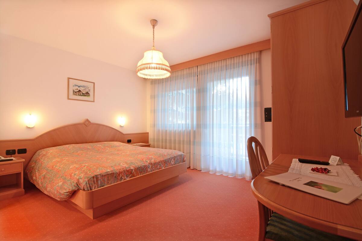 A hotel room with a bed, a desk with a laptop, a chair, a wooden wardrobe, and a window with curtains.