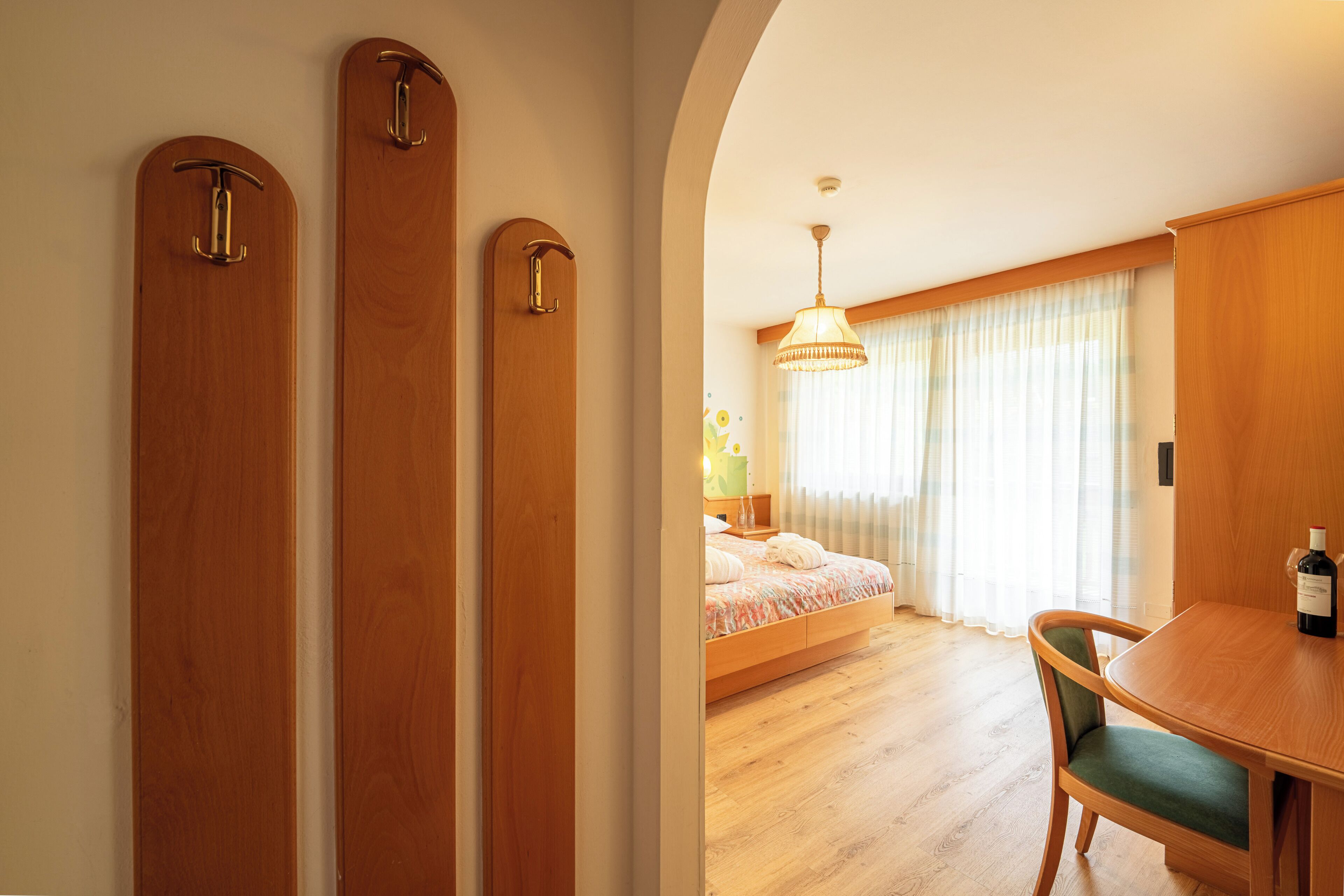 A hotel room with a wooden wardrobe, a bed with a floral bedspread, a dining table with a bottle of wine, and a chandelier.