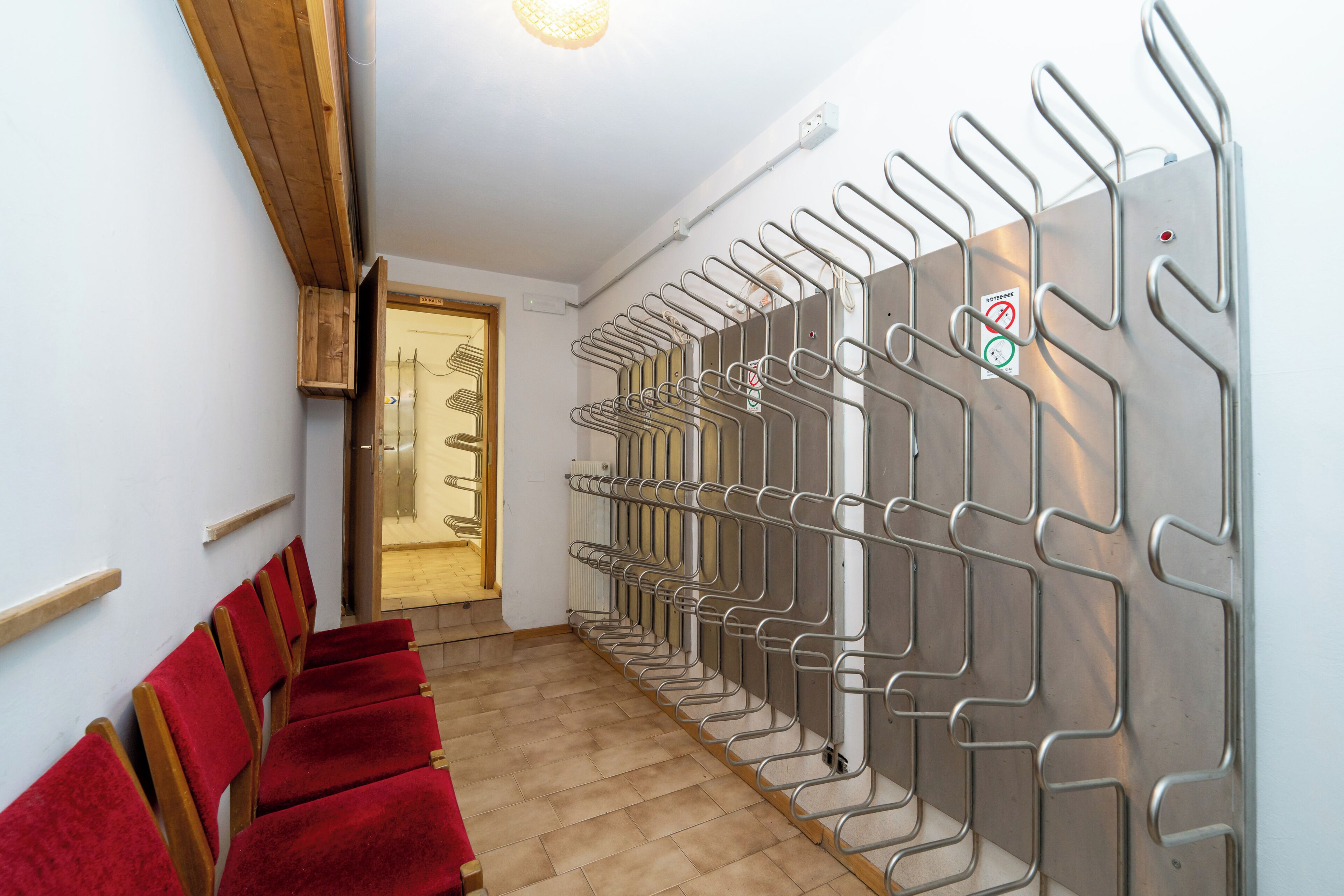 Un couloir d’hôtel avec, sur la gauche, une rangée de chaises recouvertes de tissu rouge, sur la droite, un présentoir en métal muni de crochets, et, au bout, une porte en bois.