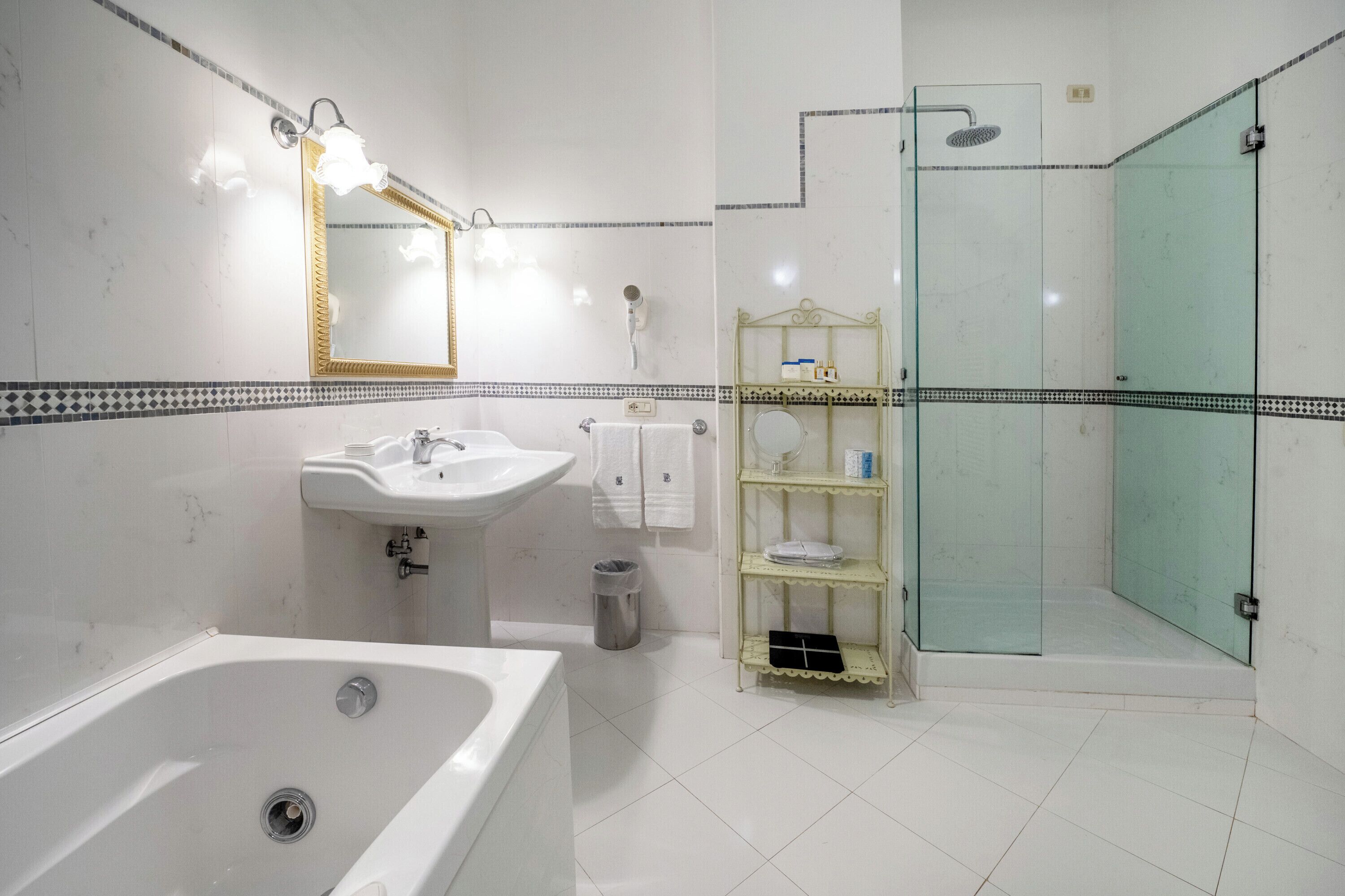 Un baño con bañera, lavamanos, espejo, cabina de ducha de vidrio y estante con artículos de aseo.