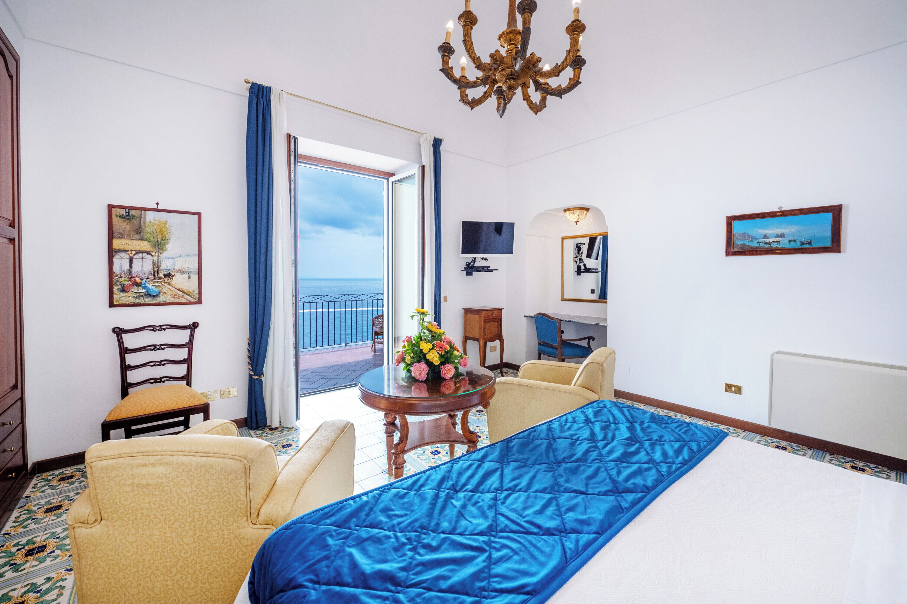 A hotel room with a bed, two armchairs, a small table with a vase of flowers, a television, and a balcony with a view.