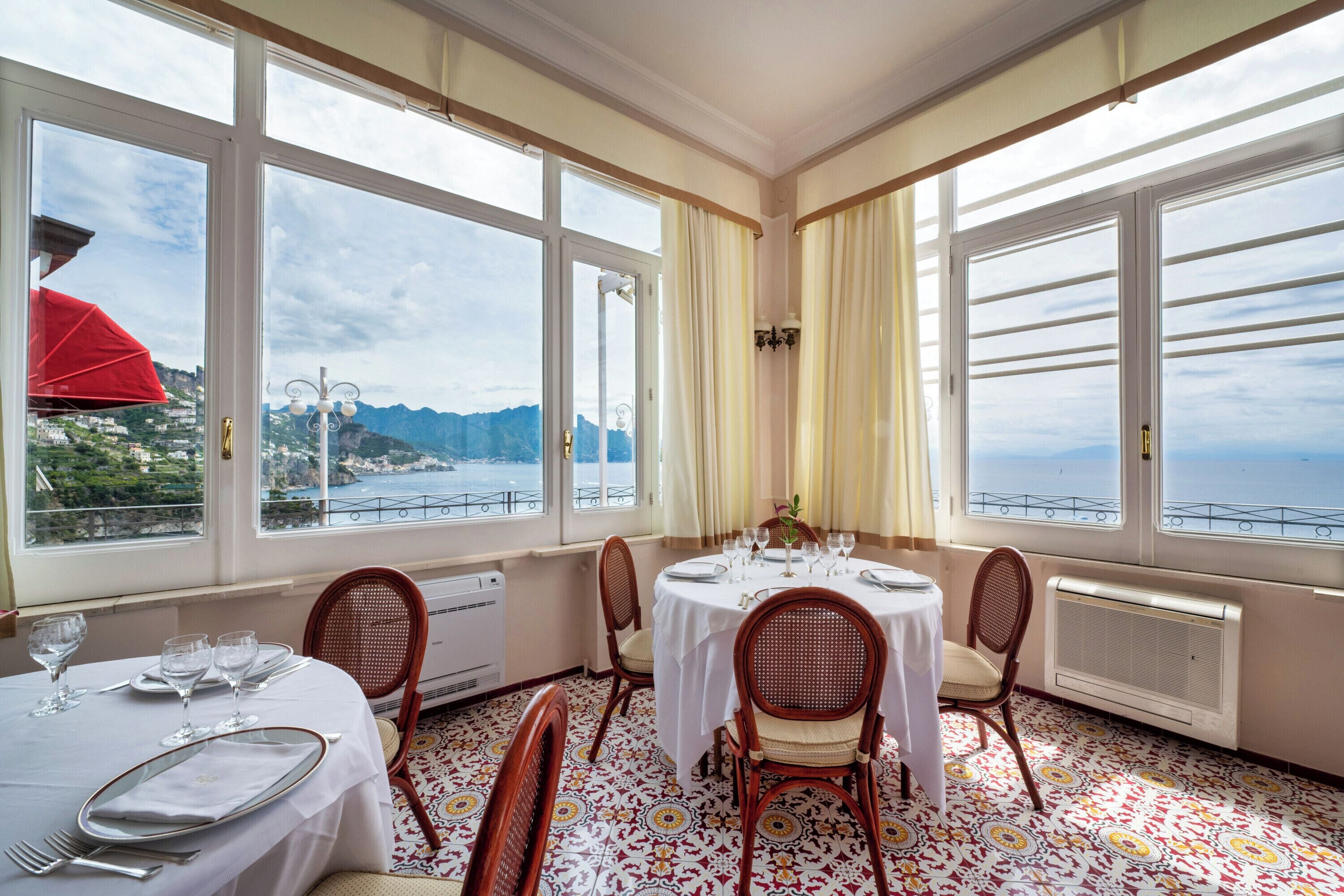 A dining area with a table set for four, large windows with curtains, and a view of the sea and mountains.