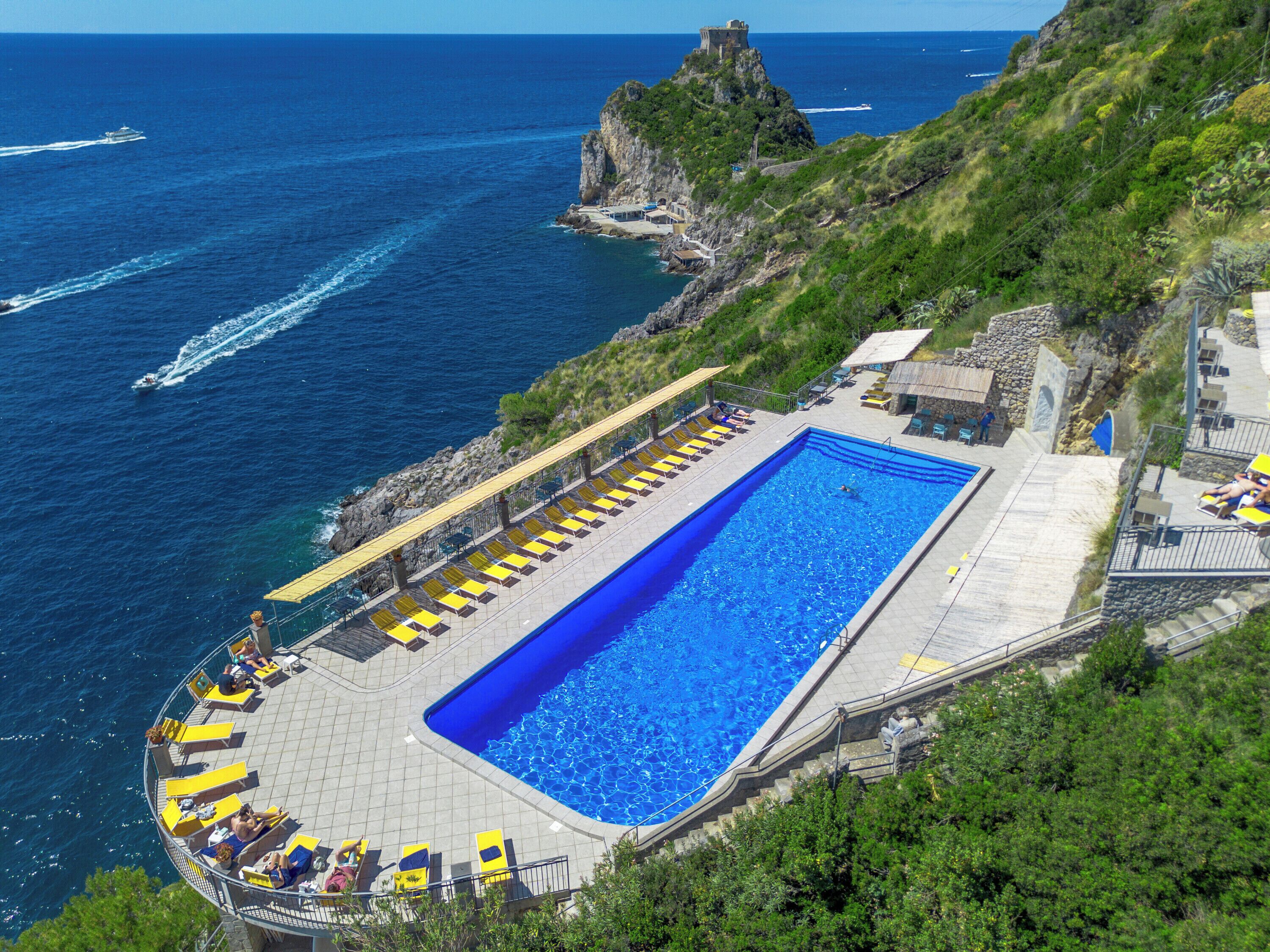 A large rectangular swimming pool with a clear blue water surface, surrounded by a tiled deck with yellow and brown lounge chairs.