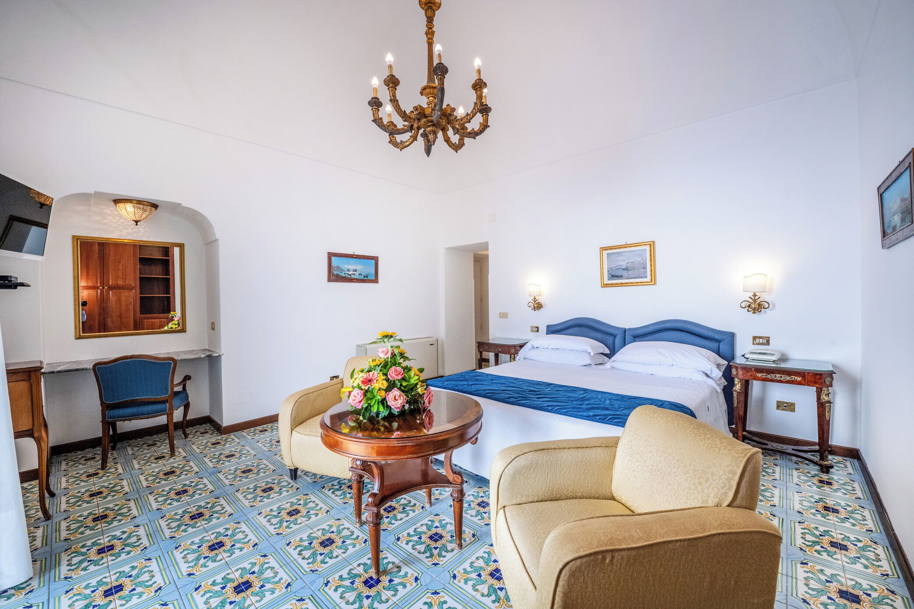A hotel room with a large bed, a desk, a chair, a small table with flowers, and a chandelier.