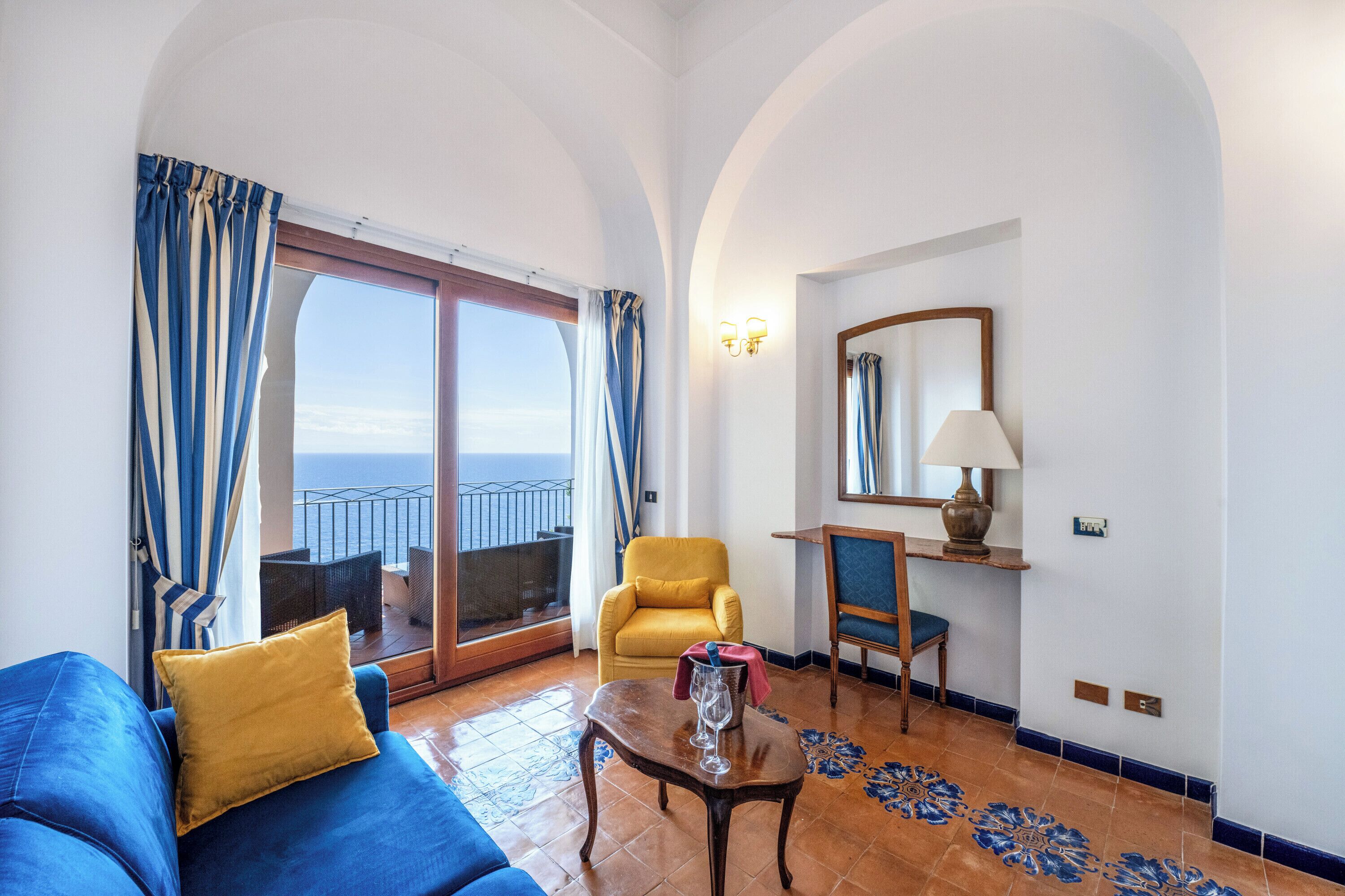 A room with a blue sofa, a yellow armchair, a small table with a glass, and a view of the sea through a sliding door.