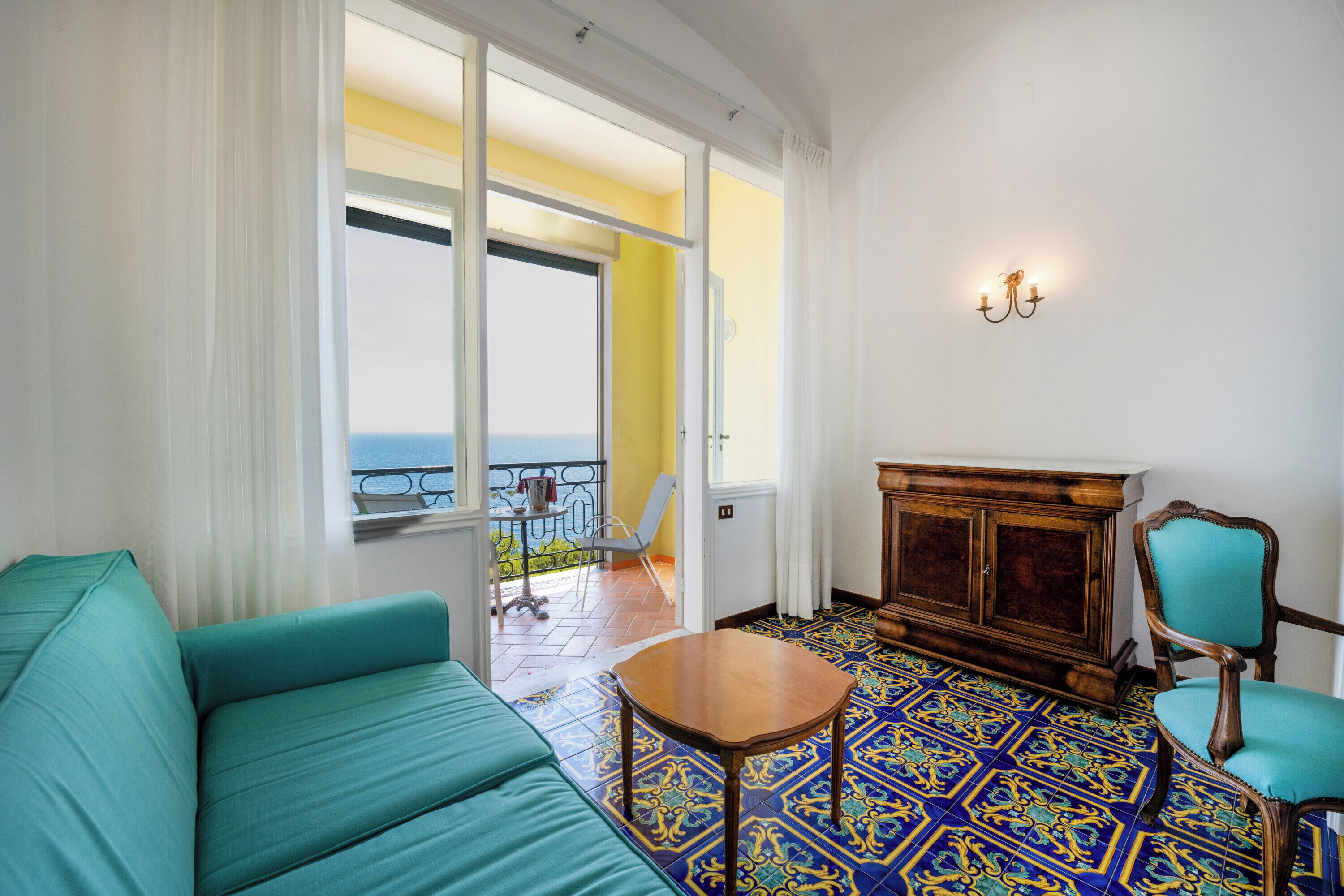 Una habitación con sofá color turquesa, un armario de madera y una silla. Hay un balcón con vistas al mar.