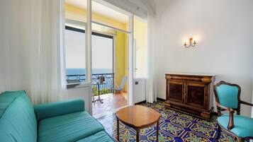 Una habitación con sofá color turquesa, un armario de madera y una silla. Hay un balcón con vistas al mar.