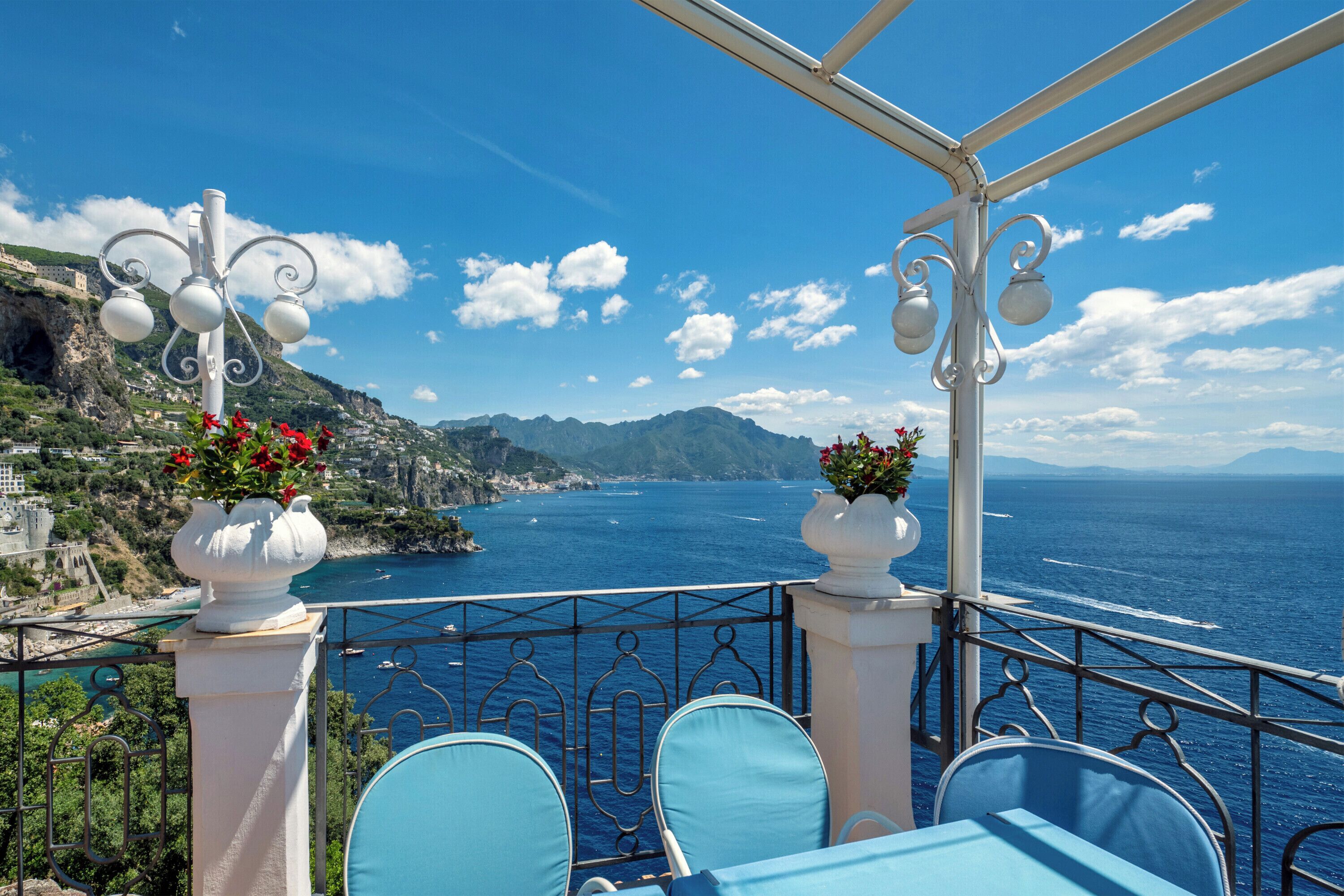 A terrace with a table and chairs, overlooking a coastal view with mountains and the sea.