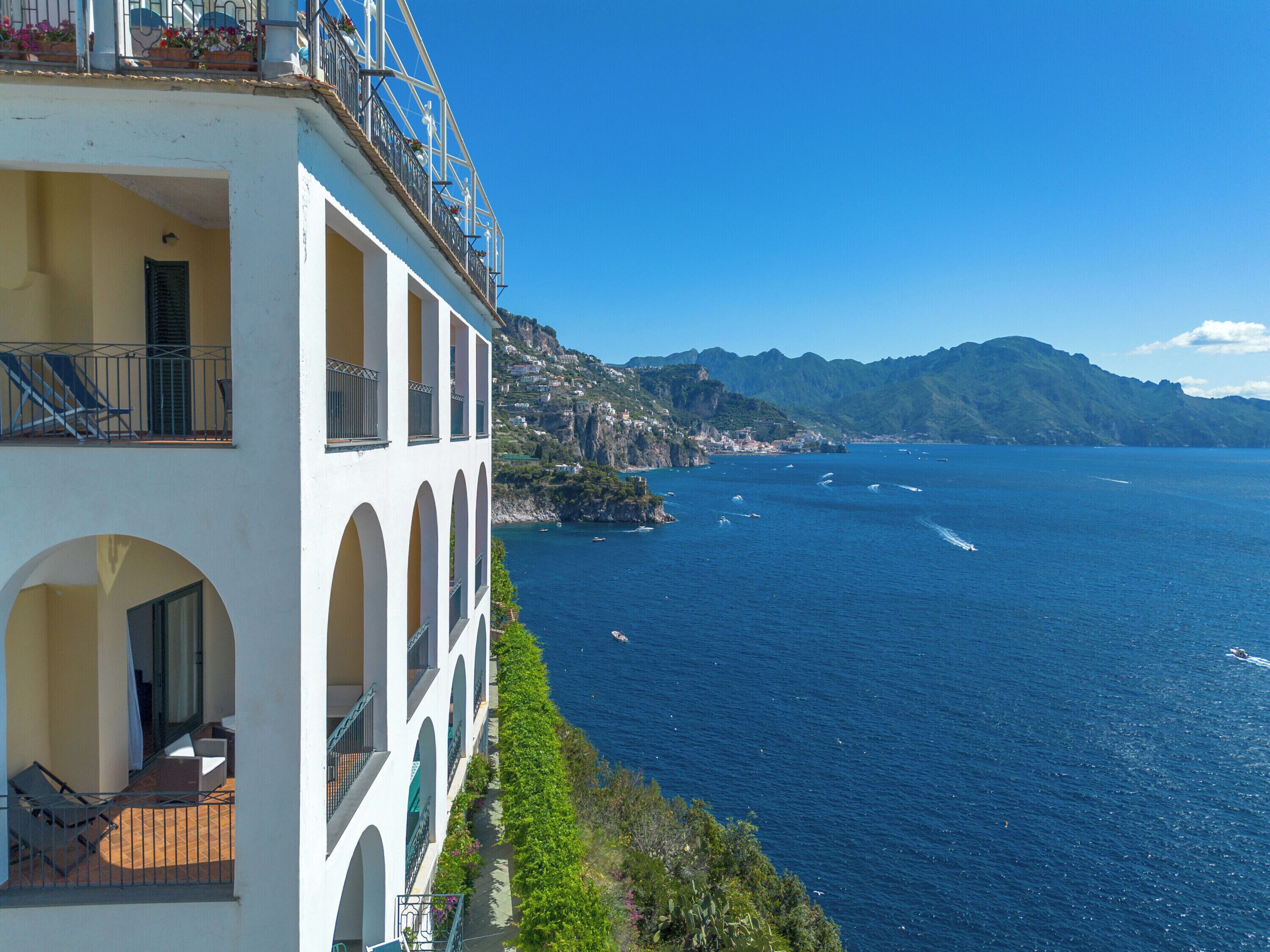 A coastal building with multiple balconies overlooking a clear blue sea and a mountainous backdrop.