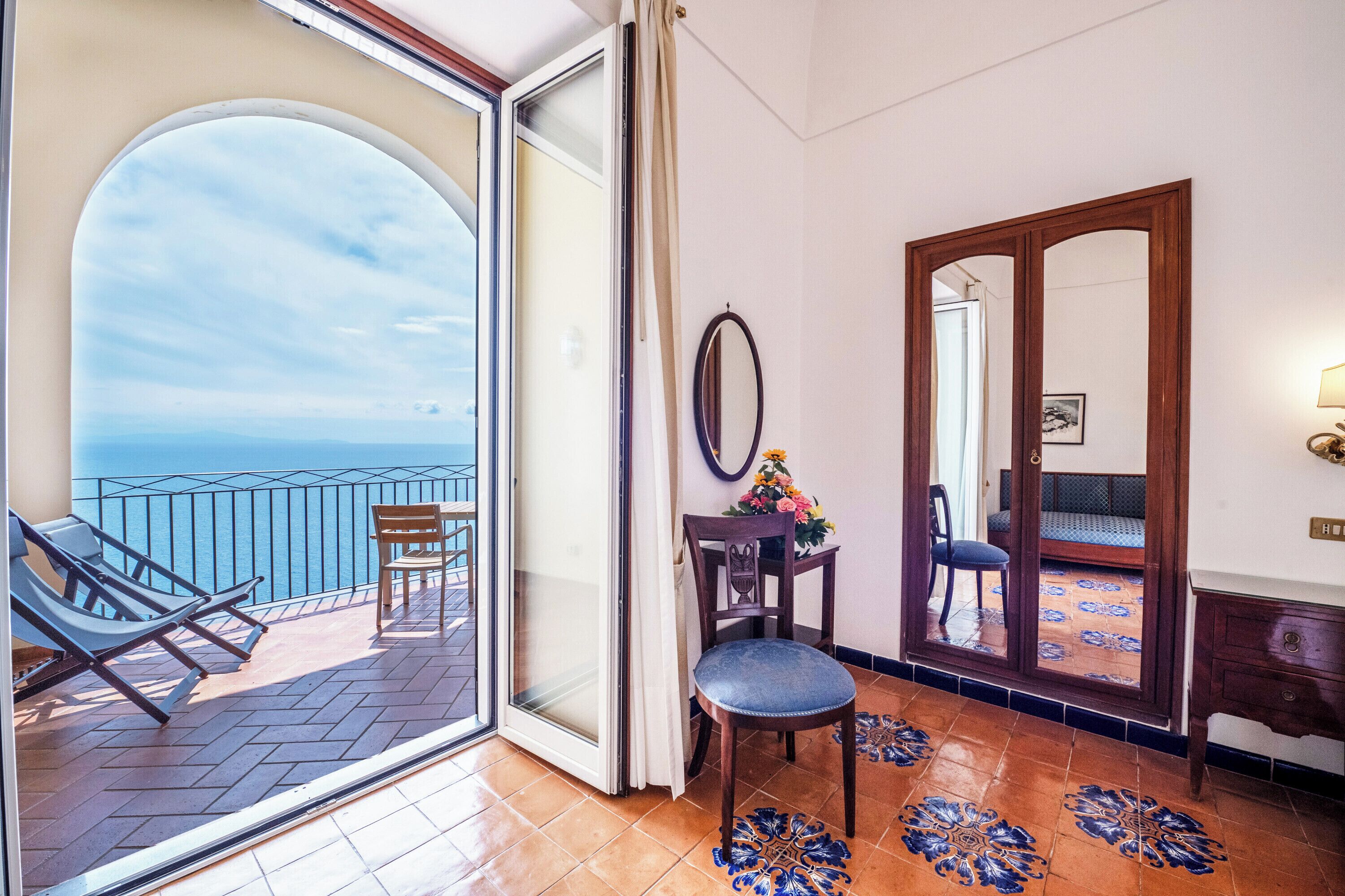 Un balcone con vista sul mare, una sedia di legno, un tavolo con un vaso di fiori e uno specchio appeso al muro.