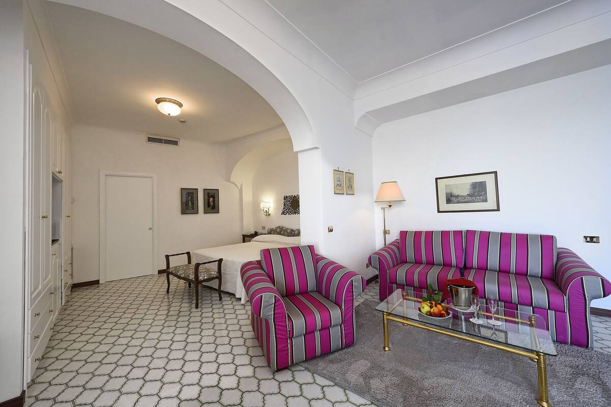 A living room with a sofa, armchair, and coffee table. There is a bed in the background and a door to the left.