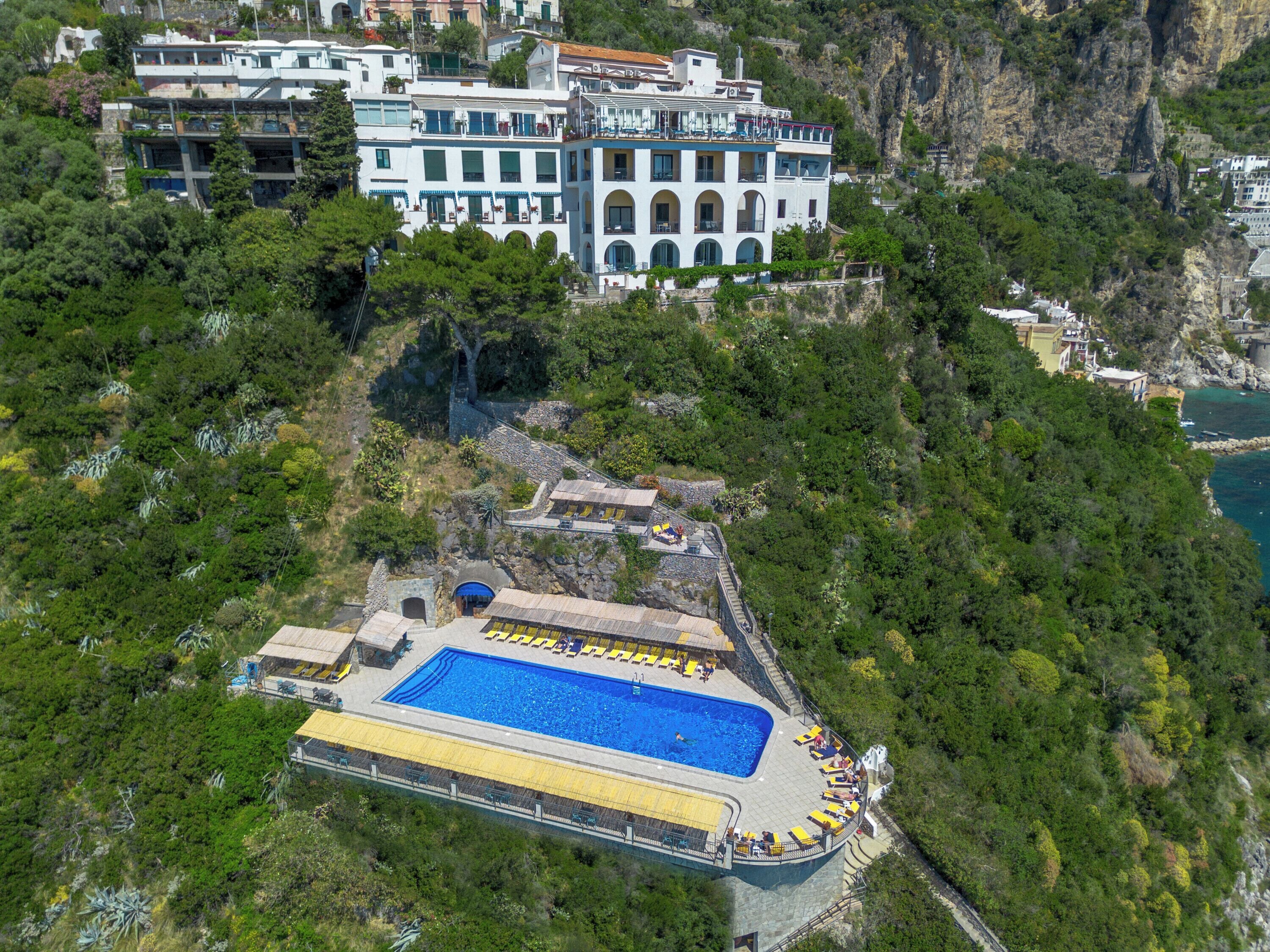 Une vue aérienne d’un hôtel doté d’une grande piscine entourée de transats, le tout se détachant sur un paysage verdoyant et des falaises rocheuses.