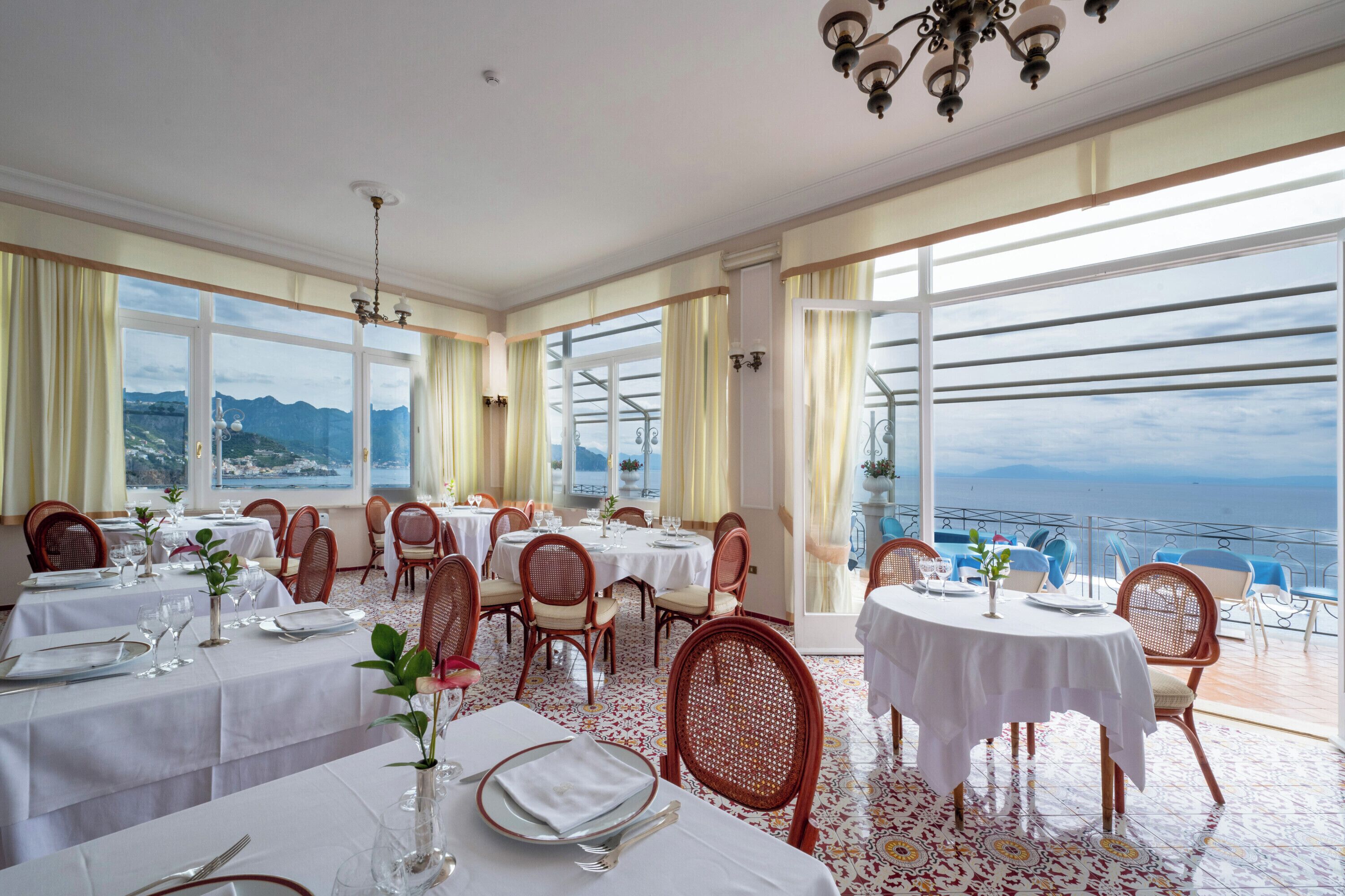 A restaurant with multiple tables and chairs, large windows with a view of the sea and mountains, and a chandelier.