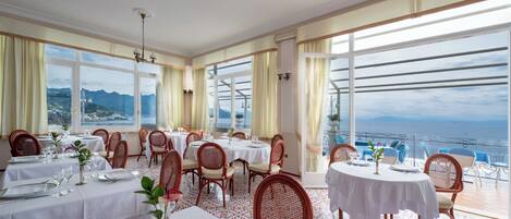 A restaurant with multiple tables and chairs, large windows with a view of the sea and mountains, and a chandelier.