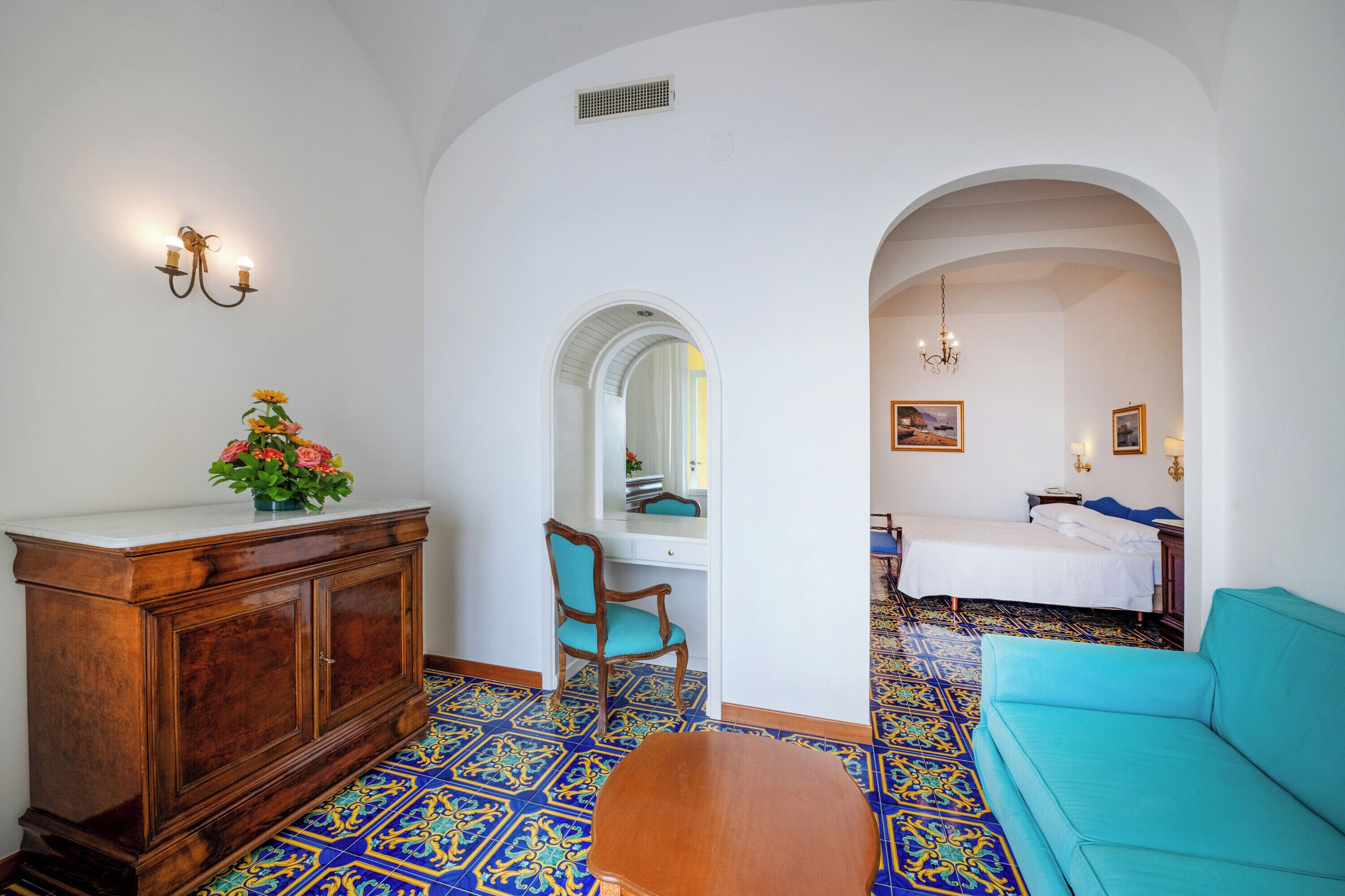 A room with a wooden cabinet, a turquoise sofa, a patterned floor, and a bed with a white bedspread.