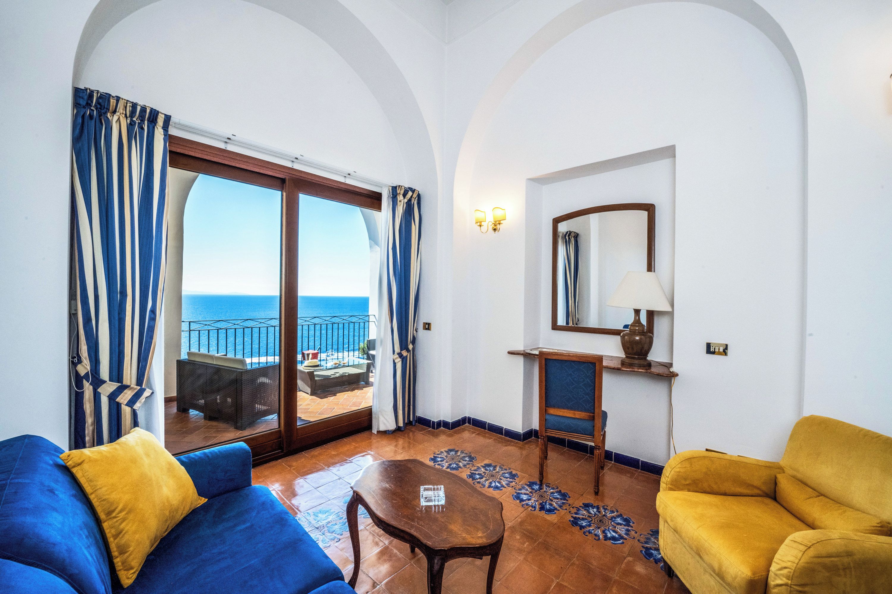 Una habitación con sofá azul, sillón amarillo, mesa de centro de madera y vista al mar a través de un ventanal corredero.
