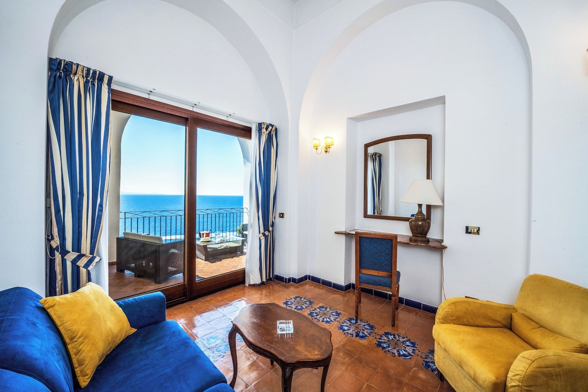A room with a blue sofa, a yellow armchair, a wooden coffee table, and a view of the sea through a sliding glass door.