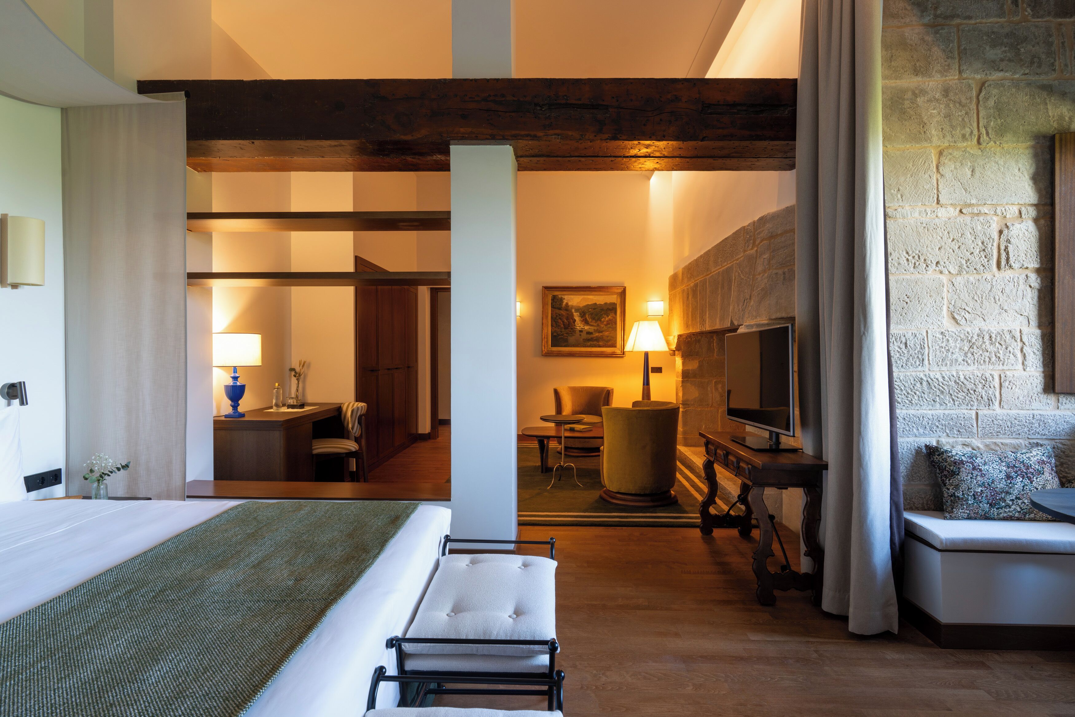 A hotel room with a bed, a desk, a TV, a sofa, and a stone wall.