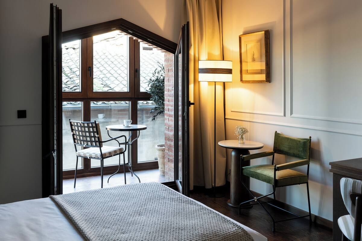 A hotel room with a bed, a small table with a chair, a lamp, and a window with curtains.