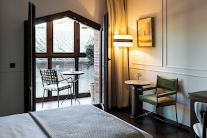 Una habitación de hotel con una cama, una mesita pequeña con una silla, una lámpara y un ventanal con cortinas.