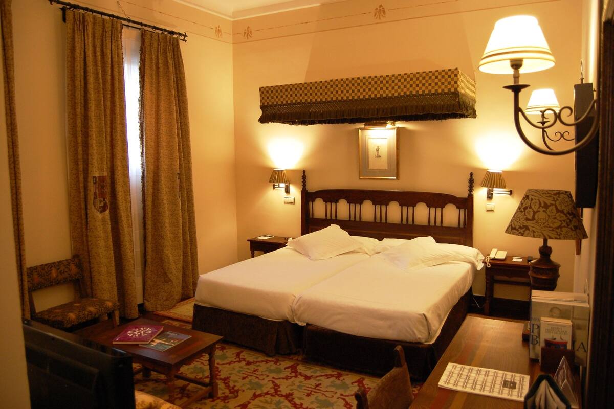 A hotel room with a double bed, two bedside lamps, a nightstand, a chair, a small table, and a patterned rug.
