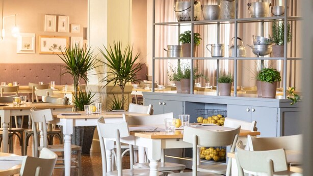 Un restaurante con sillas y mesas blancas, un estante de metal con macetas y plantas, y cuadros en la pared.