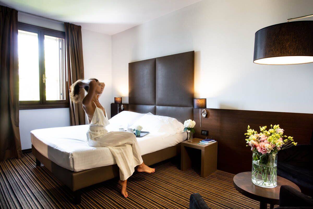 A hotel room with a large bed, a bedside table with a lamp, a small table with a vase of flowers, and a window with curtains.