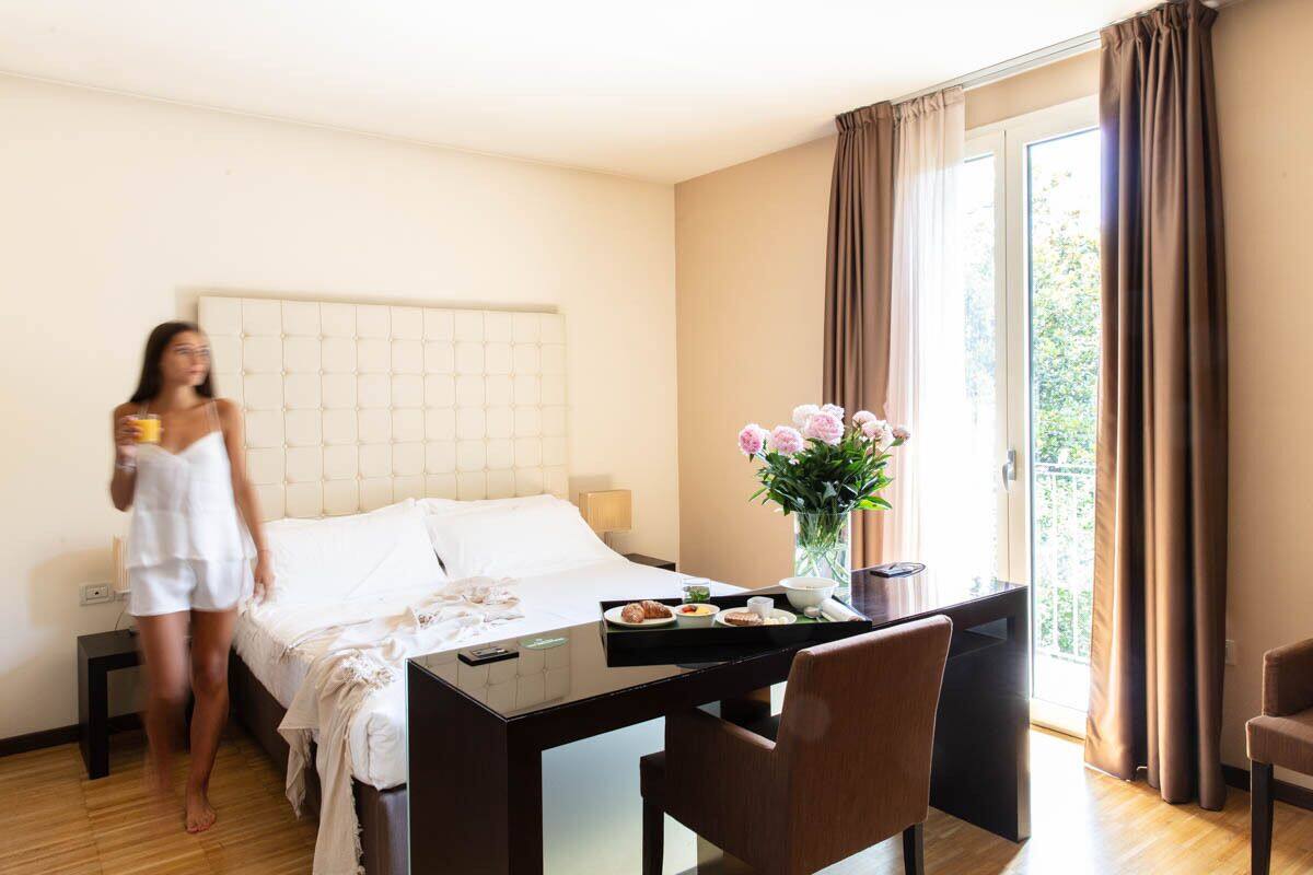 A hotel room with a bed, a dining table with breakfast, a vase with flowers, and a view of the outdoors.