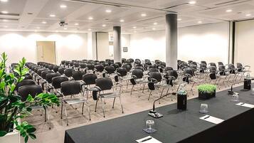 A conference room with rows of chairs, a long table with microphones, and a projector screen.