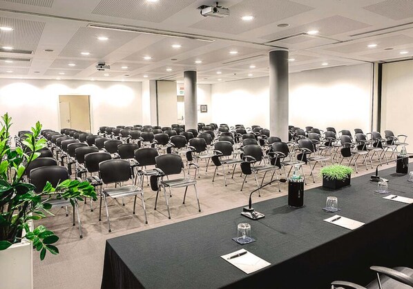Un salón de conferencias con filas de sillas, una mesa larga con micrófonos y una pantalla para proyector.