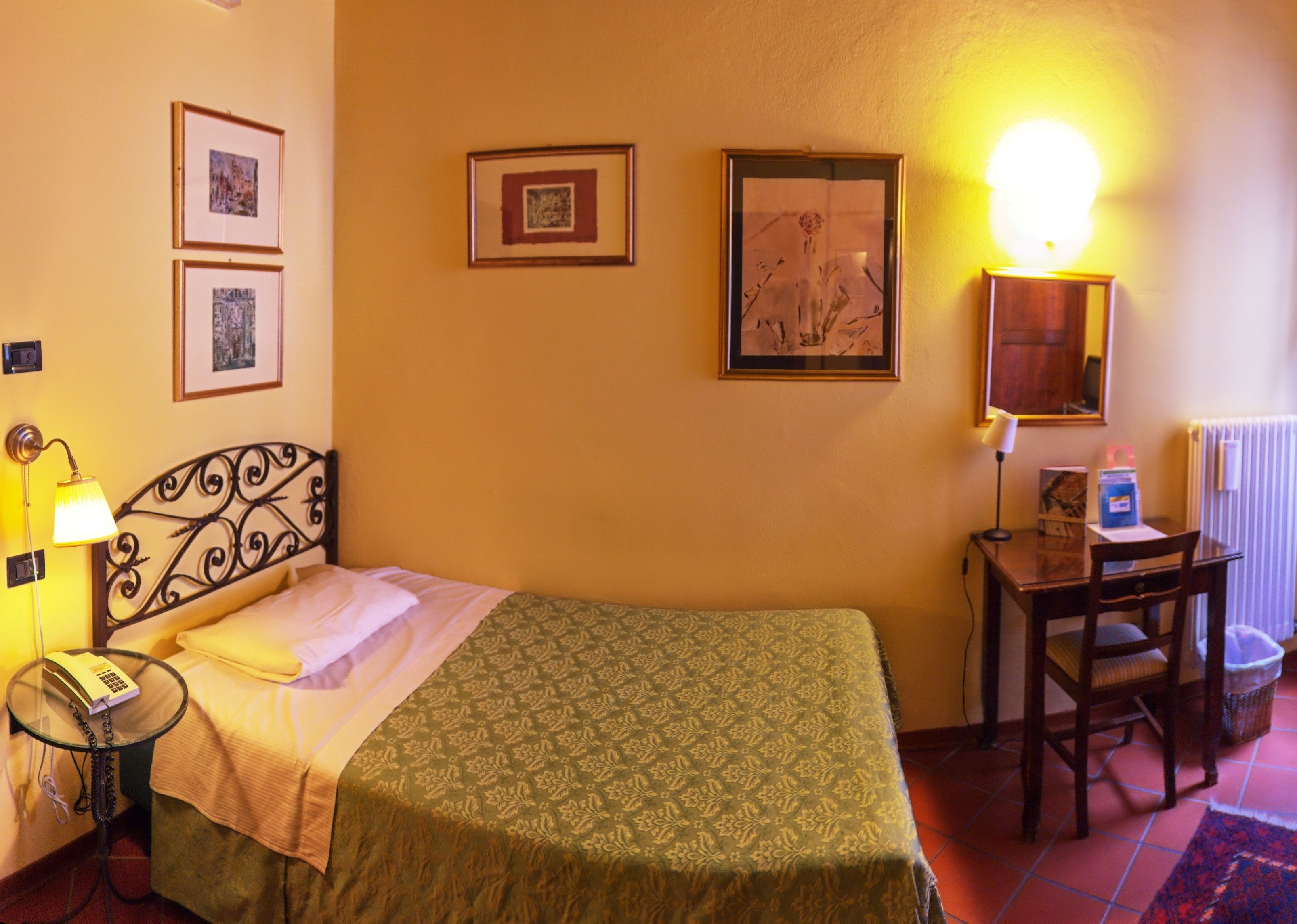 A hotel room with a bed, a desk, a chair, and a lamp. There are framed pictures on the wall.