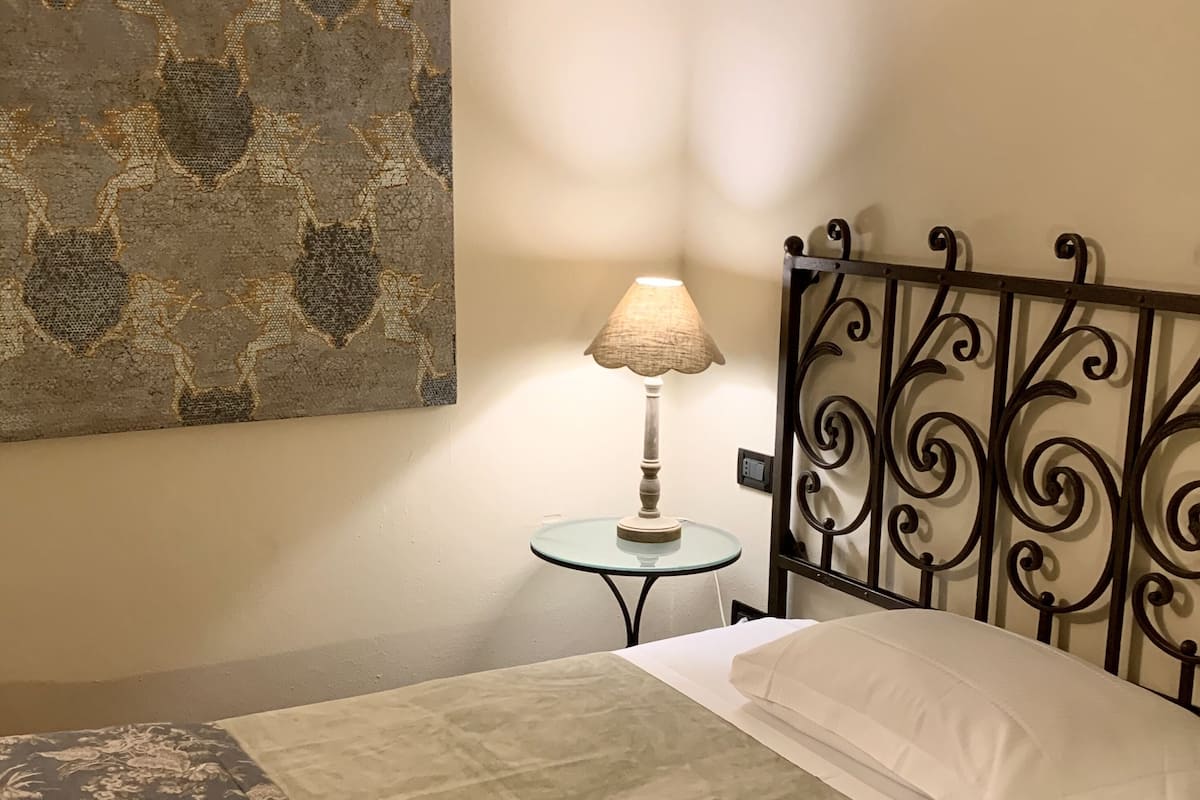 Ein ordentlich bezogenes Bett mit einem blumigem Überwurf, ein Nachttisch mit Lampe und ein dekoratives Wandbild.