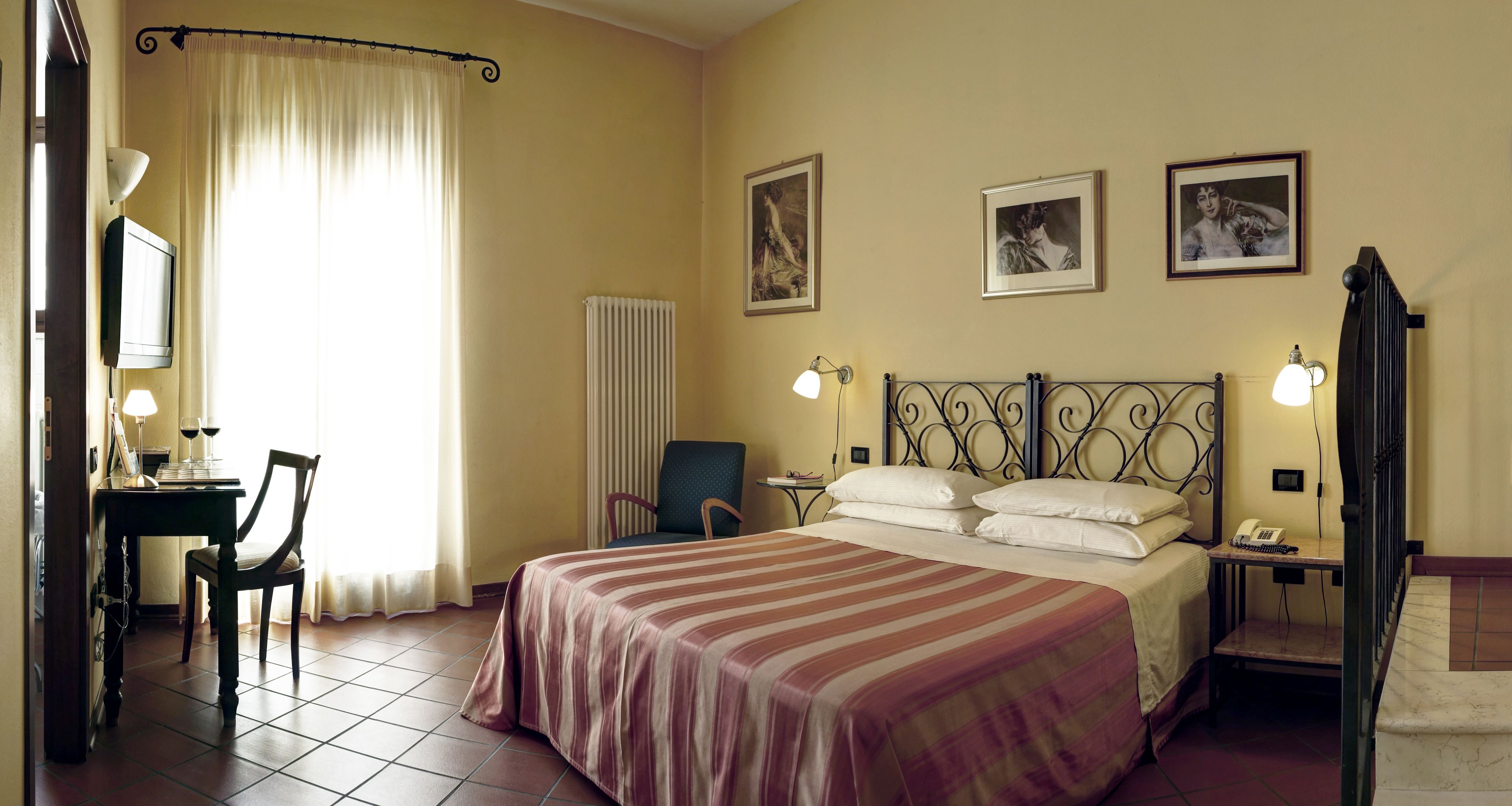 A hotel room with a bed, two chairs, a desk with a lamp, a radiator, and three framed pictures on the wall.