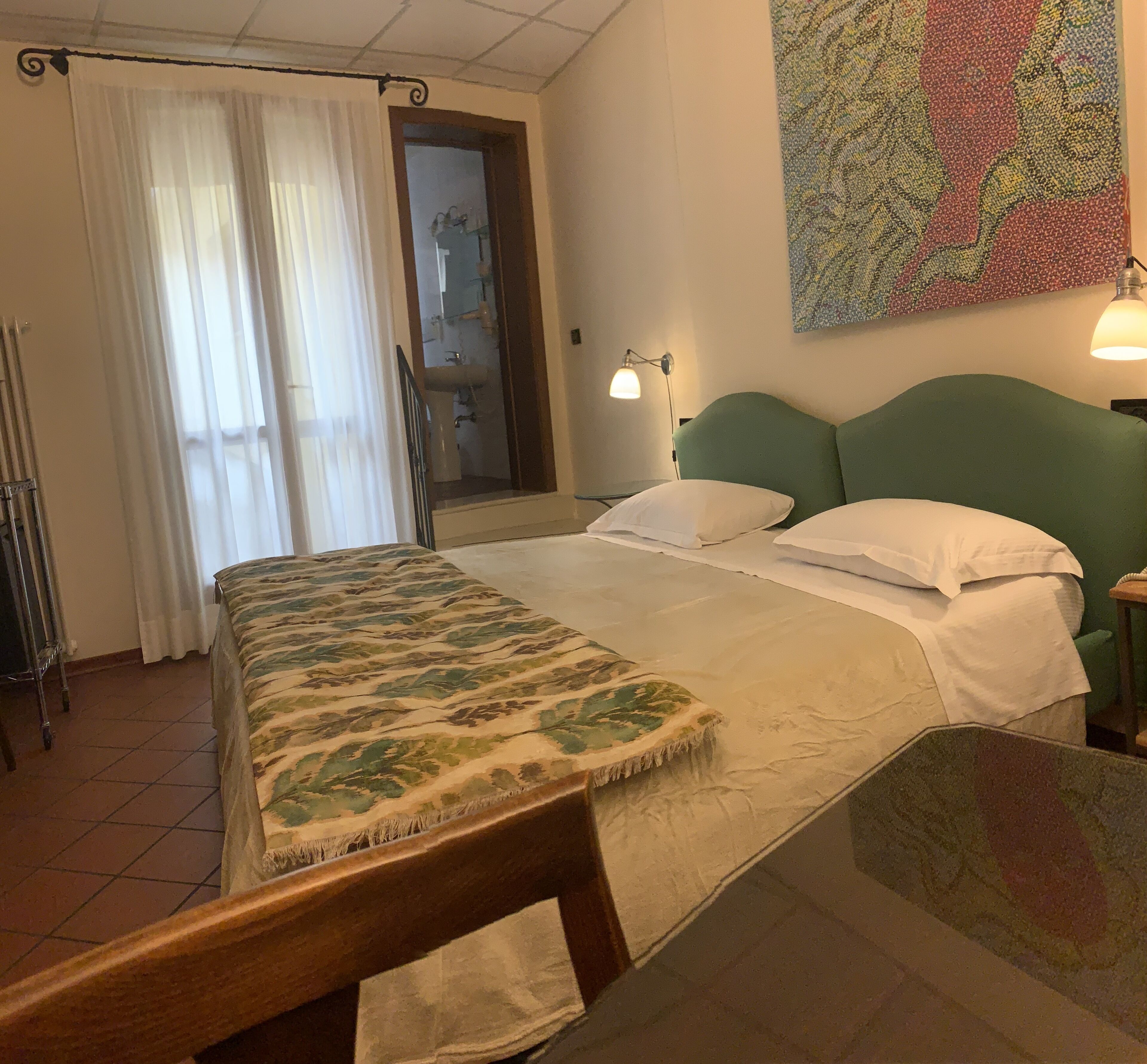 A hotel room with a bed, a nightstand, a lamp, a window with curtains, a radiator, and a painting on the wall.