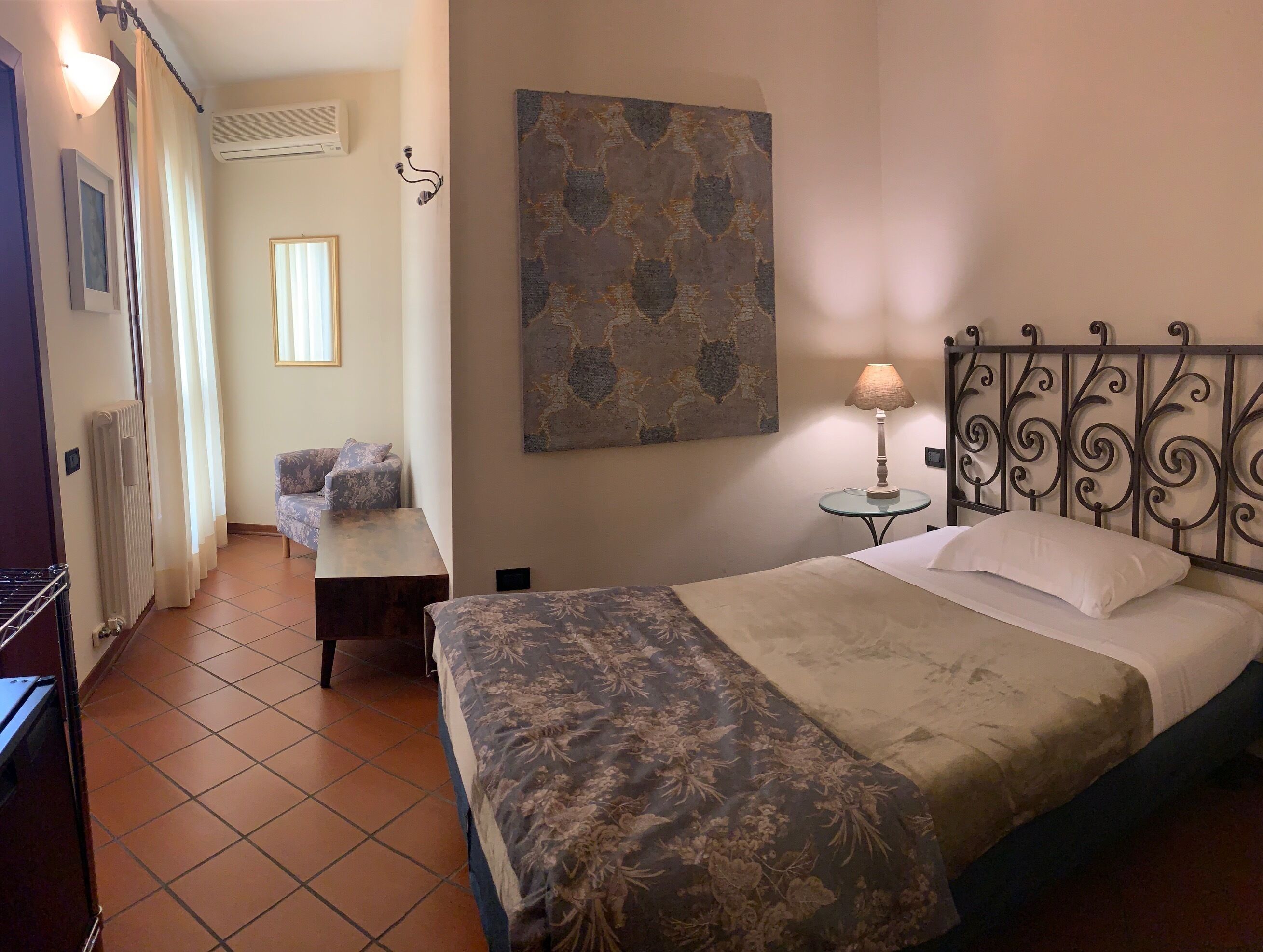 A bedroom with a bed, a bedside table with a lamp, a chair, a radiator, and a painting on the wall.