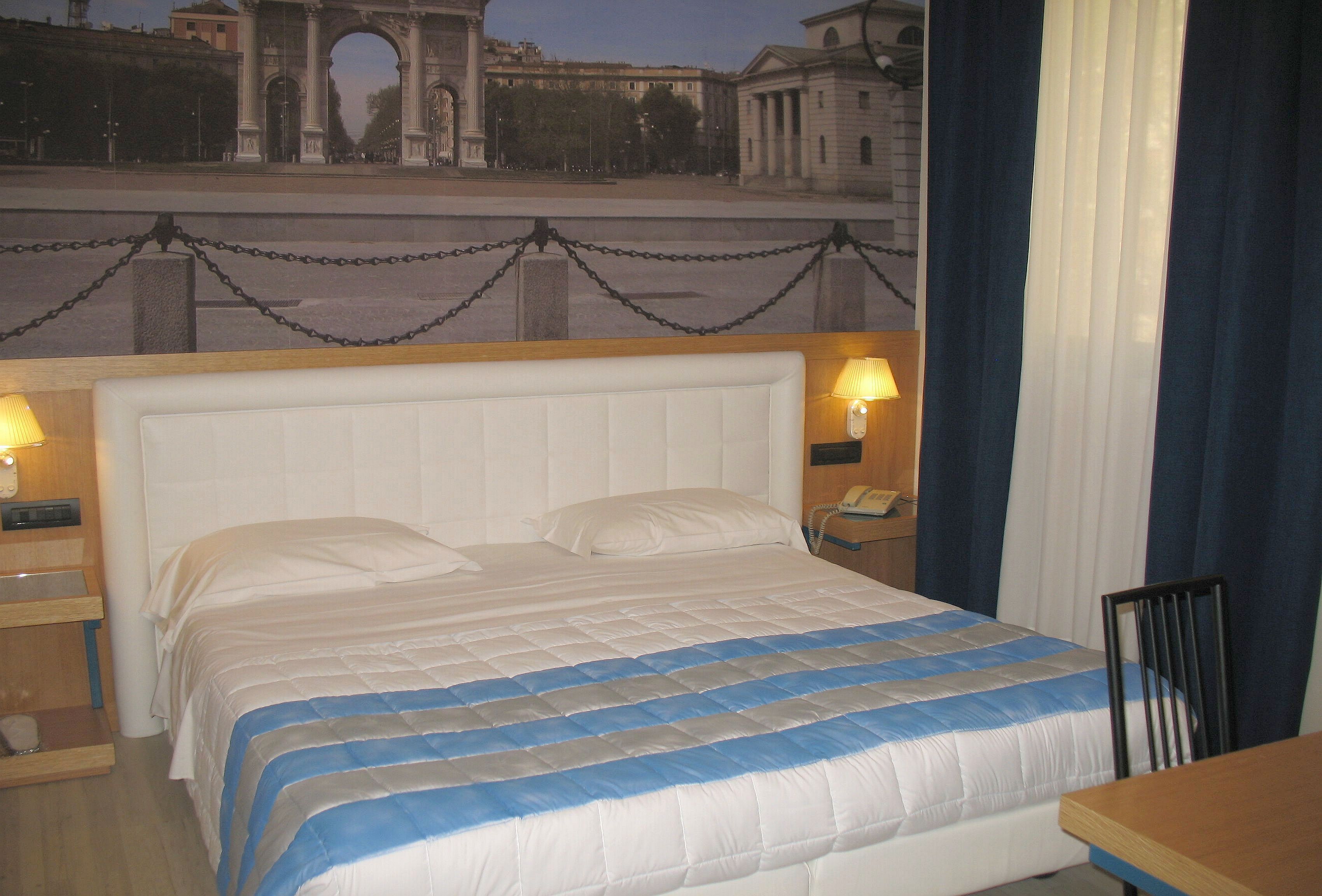 A hotel room with a bed, bedside lamps, a nightstand, a wooden table, and a view of a cityscape with an archway.