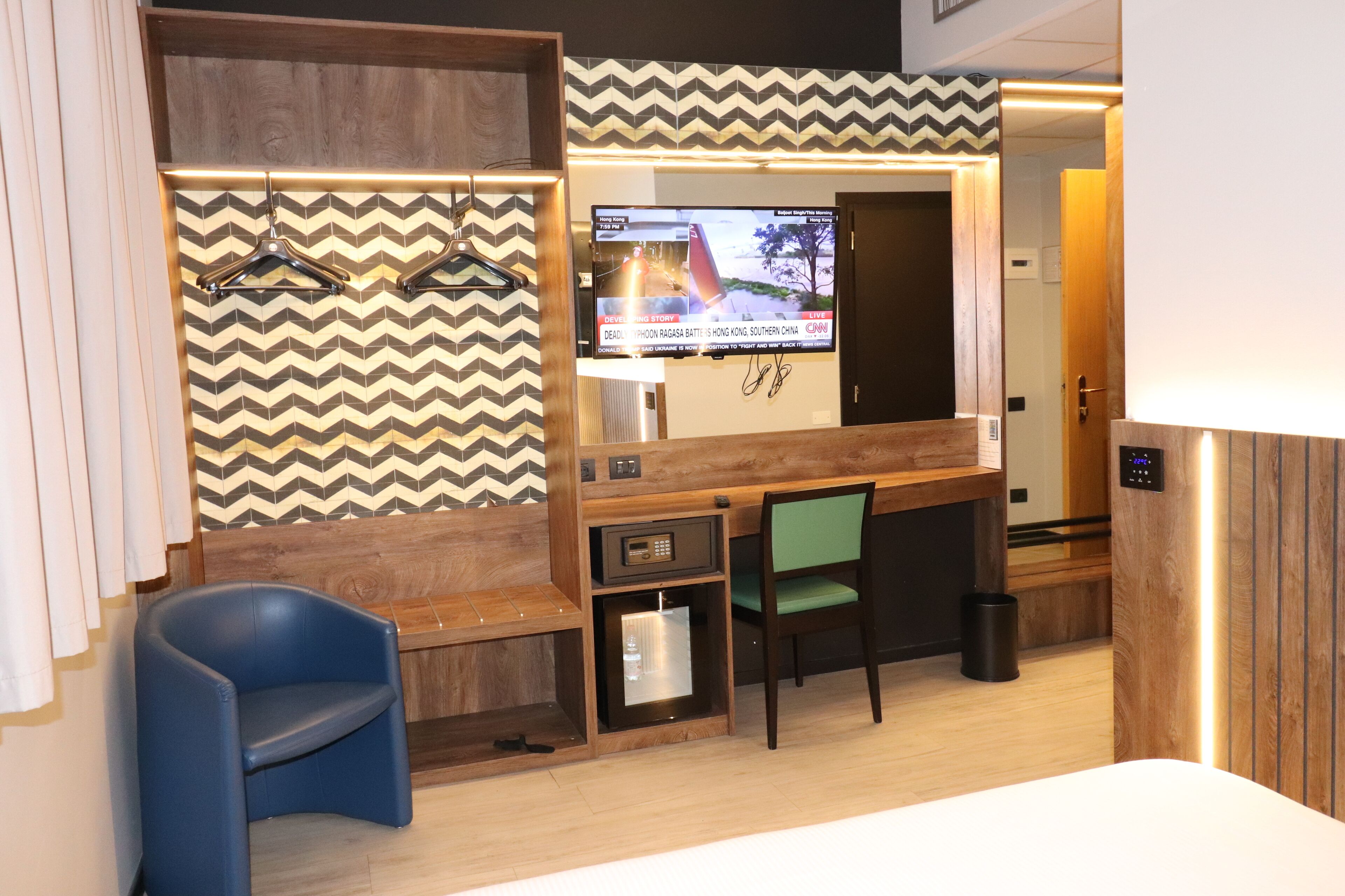 A hotel room with a wooden bench, a blue chair, a desk with a computer, and a television displaying a news channel.